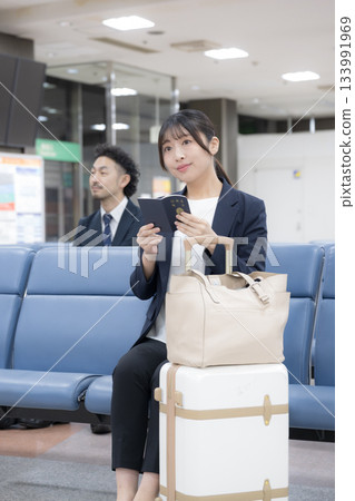 一位商務女性坐在候機大樓裡，手裡拿著護照。這是在機場或車站候機大樓的商務旅行場景。全身照。 133991969