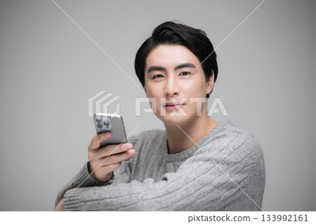 一位英俊的男子正在看智慧型手機，背景是灰色的，他看著鏡頭，面帶微笑。 133992161