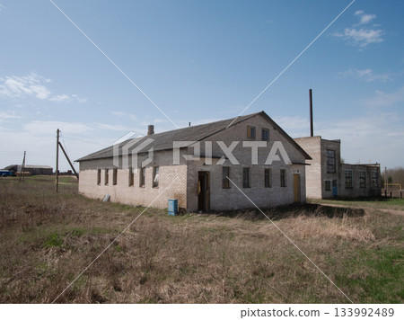 A weathered brick building stands alone in a vast grassy field under a bright blue sky with scattered clouds, showcasing signs of neglect and abandonment. A weathered brick building stands alone in a vast grassy field under a bright blue sky with scattered clouds, showcasing signs of neglect and abandonment. 133992489