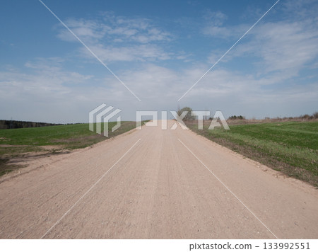 A long, dusty dirt road leads through vibrant green fields under a bright blue sky with a few clouds, capturing the tranquility of rural life on a clear day. 133992551