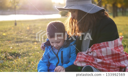 A mother and her young son sit together in a park, enjoying the sunshine and exploring the grass as autumn leaves decorate the surroundings, creating a cozy atmosphere. A mother and her young son sit together in a park, enjoying the sunshine and exploring the grass as autumn leaves decorate the surroundings, creating a cozy atmosphere. 133992569
