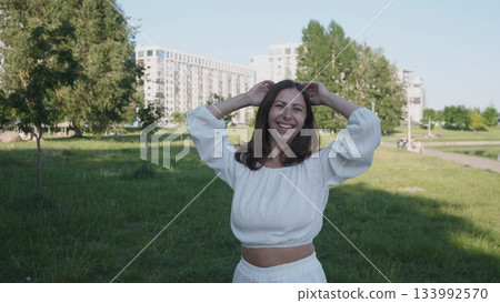 Woman wearing a white outfit stands in an urban park, smiling and enjoying the sunny weather while her hair blows in the gentle breeze. Woman wearing a white outfit stands in an urban park, smiling and enjoying the sunny weather while her hair blows in the gentle breeze. 133992570