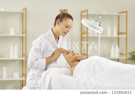 Young female cosmetologist examining woman client skin condition in beauty salon. Young female cosmetologist examining woman client skin condition in beauty salon. 133992651