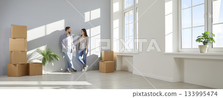 Happy young couple standing against the wall with cardboard boxes in a new apartment on moving day. 133995474