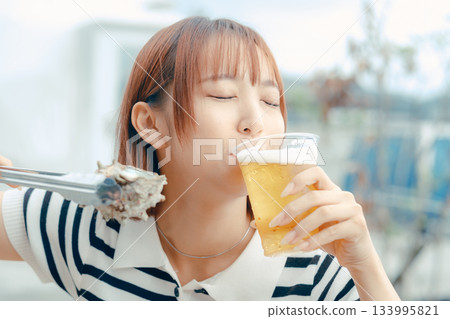 一位二十多歲的女子一邊享用啤酒,一邊燒烤海鮮。 一位二十多歲的女子一邊享用啤酒,一邊燒烤海鮮。 133995821