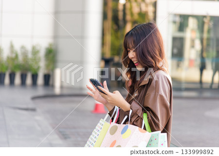 Asian woman enjoys shopping, active lifestyle for people, holding fashion bags near mall entrance, smiling brightly, posing outdoors after black friday shop event, checking mobile phone. Asian woman enjoys shopping, active lifestyle for people, holding fashion bags near mall entrance, smiling brightly, posing outdoors after black friday shop event, checking mobile phone. 133999892