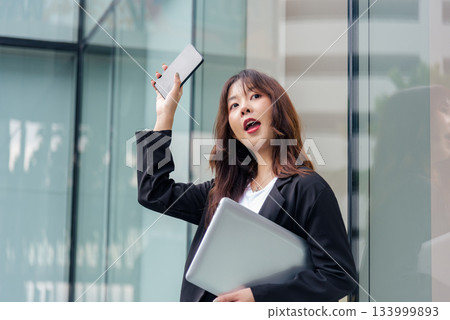 Young Asian business woman wearing suit, ready for work, holding laptop, raising phone with open mouth expression on busy city street, urban office building backdrop shows dynamic professional life. 133999893