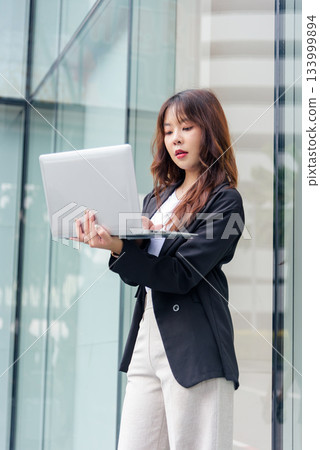 Young Asian woman in business suit works on street, busy professional holding laptop, looking intently at screen in front of modern building glass exterior. 133999894