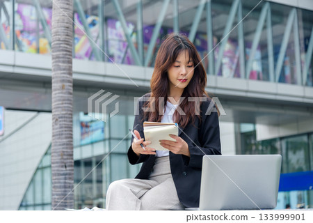 Young Asian woman, busy with business work, wears suit, sits on street, writes in notebook, laptop nearby, embodying professional modern urban lifestyle, dedicated to her tasks in bustling city. Young Asian woman, busy with business work, wears suit, sits on street, writes in notebook, laptop nearby, embodying professional modern urban lifestyle, dedicated to her tasks in bustling city. 133999901