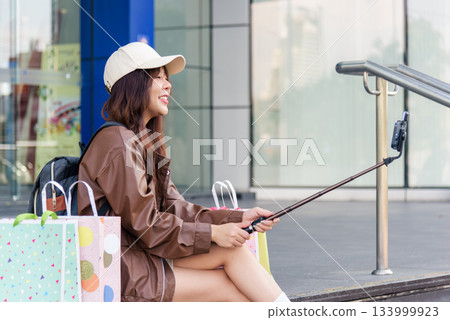 Asian woman enjoys shopping lifestyle, posing with colorful bags near modern mall, holding phone for memory, fashion conscious people find retail happiness. 133999923