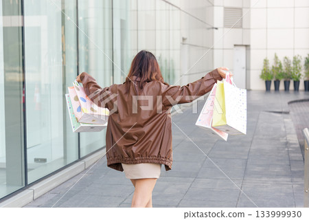 Asian woman enjoys shopping lifestyle, holding numerous colorful fashion bags with arms outstretched, rear view shows people engaging in retail therapy outside modern city mall environment. 133999930