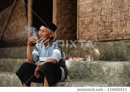 An elderly man is quietly sitting and smoking on the steps of a traditional home 134000913