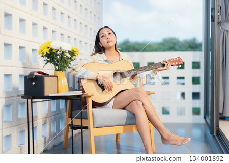 A young woman plays a guitar on a couch in her living room near her porch. On a weekend, a young woman enjoys herself. People, Lifestyle, Hobbies, Home Life.	 134001892