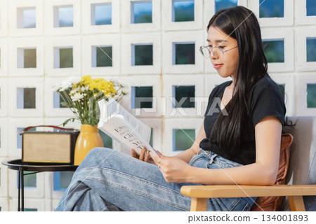 A young Asian woman reads a book on a couch on her porch at home on a weekend without electronics. The concept of digital detox and social media. Reading is a leisure activity.	 134001893