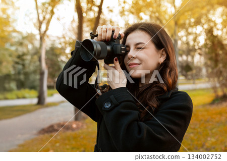 Young woman reviewing photos on her digital camera while standing in a sunny park. Warm autumn light, creative outdoor photography moment, lifestyle scene. 134002752