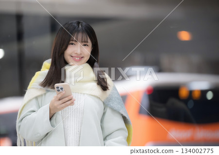 A woman wearing a cute scarf and holding a smartphone at a place that looks like a platform or bus terminal at a winter train station 134002775