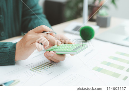 Close-up of hand using green calculator on financial charts with eco earth globe, symbolizing sustainable finance, green business planning, environmental accounting and clean economic growth. 134003337