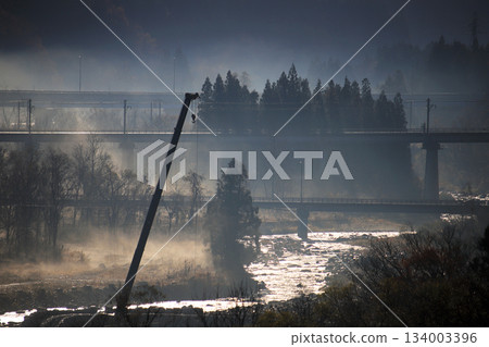 A misty mountain river, a construction crane and coniferous trees, and an overpass and highway in the distance. 134003396