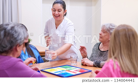 Caregiver serving water to senior women playing board game 134004441