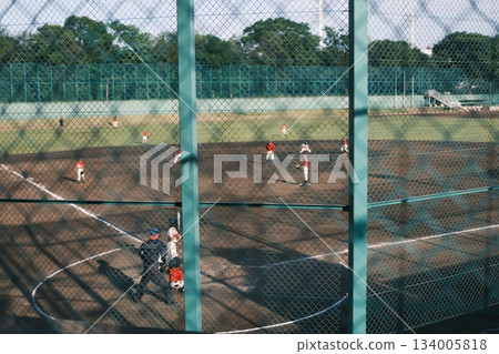 A hotly contested game of youth. A summer baseball field seen through the wire fence A hotly contested game of youth. A summer baseball field seen through the wire fence 134005818
