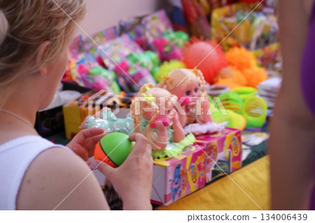 The photograph shows a woman choosing a toy at a market or store. She's holding a green toy, and various children's toys, including dolls and other items, are laid out on the table in front of her. Ot The photograph shows a woman choosing a toy at a market or store. She's holding a green toy, and various children's toys, including dolls and other items, are laid out on the table in front of her. Ot 134006439