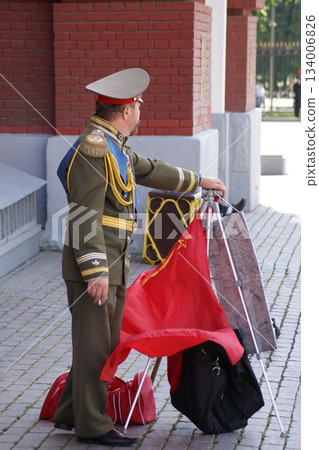 The photograph depicts a man in military uniform standing on the street, holding a red flag with a star and a hammer and sickle. Next to him is a black suitcase. The man is wearing a green uniform wit The photograph depicts a man in military uniform standing on the street, holding a red flag with a star and a hammer and sickle. Next to him is a black suitcase. The man is wearing a green uniform wit 134006826