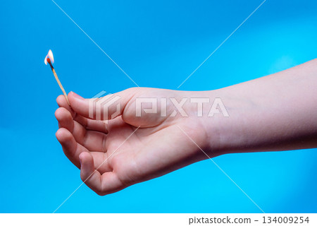 A close-up of a child's hand holding a burning match against a plain blue background. Fire safety concept. A close-up of a child's hand holding a burning match against a plain blue background. Fire safety concept. 134009254