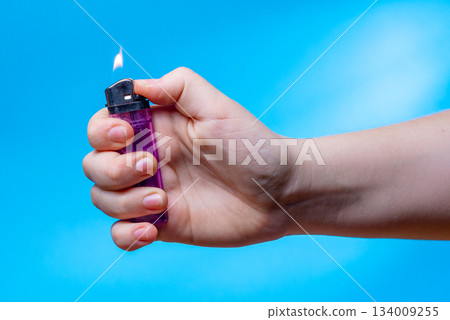 A close-up of a child's hand holding a lit lighter against a plain blue background. Fire safety concept. A close-up of a child's hand holding a lit lighter against a plain blue background. Fire safety concept. 134009255