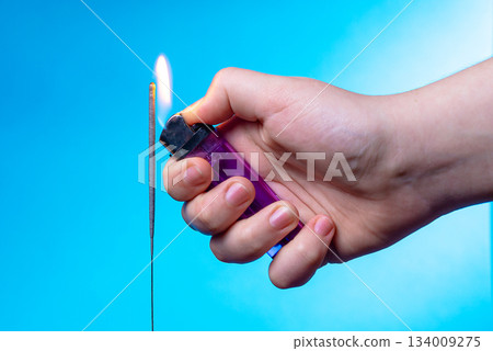 A close-up of a child's hand holding a lighter against a blue background. She's using the lighter to light a sparkler. Fire safety concept. A close-up of a child's hand holding a lighter against a blue background. She's using the lighter to light a sparkler. Fire safety concept. 134009275