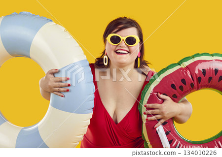 Smiling fat woman in swimsuit, sunglasses and earrings holding two inflatable beach rings Smiling fat woman in swimsuit, sunglasses and earrings holding two inflatable beach rings 134010226