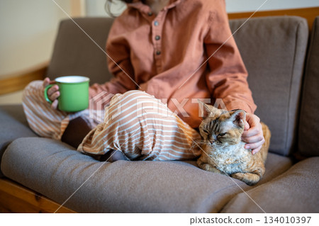 Woman in pyjamas petting domestic cat sitting on couch at home drinking tea. 134010397