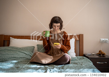 Pensive teenager sitting on bed with cup of tea, reading news in internet, hanging in social network Pensive teenager sitting on bed with cup of tea, reading news in internet, hanging in social network 134010402