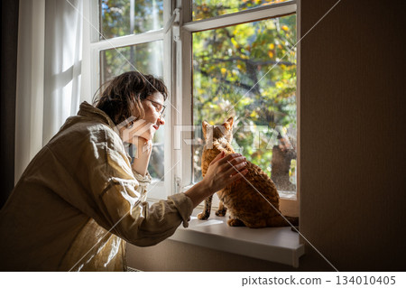 Tender pet lover stroking beloved adorable breed cat Devon Rex sitting on windowsill in sunlight 134010405
