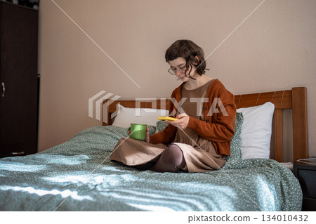 Teen girl reading messages scrolling social media in smartphone drinking tea in bed. Phone addiction 134010432