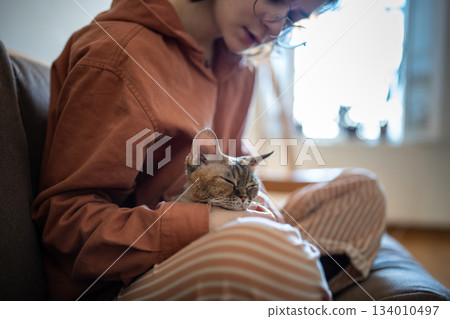 Pet lover sitting on couch, holding sleepy cat on knees. Tender friendship between person and pet Pet lover sitting on couch, holding sleepy cat on knees. Tender friendship between person and pet 134010497