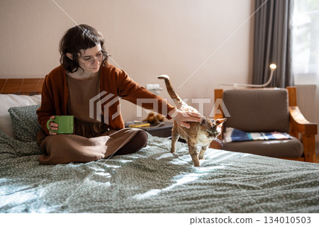 Loving teenager sitting on bed, drinking tea, caressing, stroking beloved short-haired cat Devon Rex 134010503