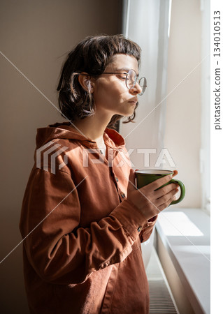 Teen girl nerd in glasses looking at window drinking cup of tea at home. Introvert, friendless 134010513