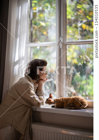 Depressed woman thinking about troubles sadly looking at window curious cat trying support owner.  134010535