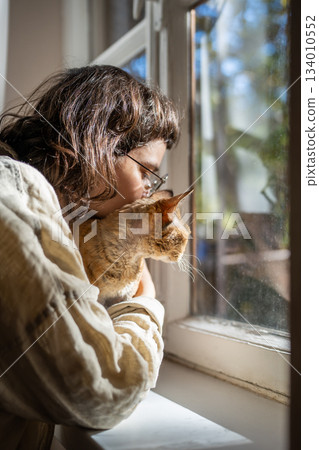 Caring pet lover cuddling beloved breed cat Devon Rex sitting on windowsill looking at window Caring pet lover cuddling beloved breed cat Devon Rex sitting on windowsill looking at window 134010552