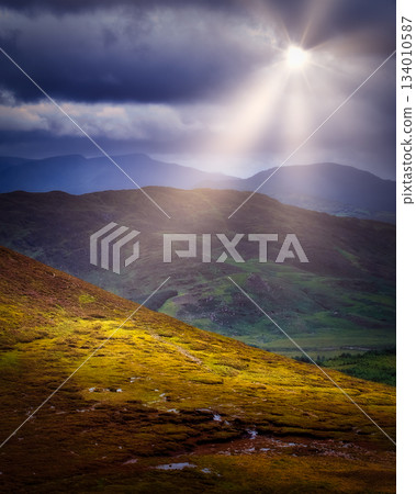 A scenic view of rolling hills and mountains under a dramatic sky. A bright ray of sunshine breaks through dark clouds, illuminating the landscape in County Kerry. 134010587