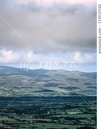 A distant view shows a valley with fields and towns below a mountain range. Clouds fill the sky above the landscape near the Coomloughra Horseshoe hiking trail. 134010588