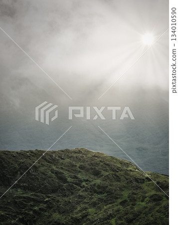 A rugged, green and rocky summit and a bright sun shining through the clouds, seen from the Horseshoe trail in Coomloughra, Ireland. 134010590