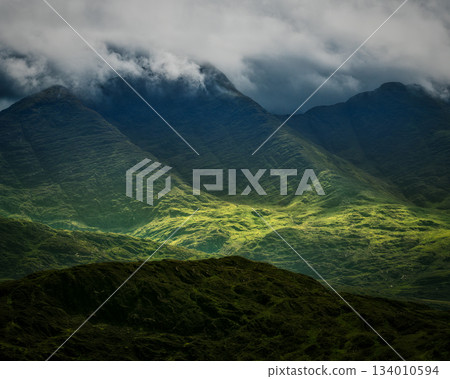 A beautiful view of green mountains shrouded in clouds during a hike along the Coomloughra Horseshoe trail. Light shines on the lower valleys creating a stunning contrast. 134010594