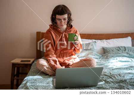 Girl watching series movie on laptop sits in bed drink tea at home leisurely passing time on day off 134010602