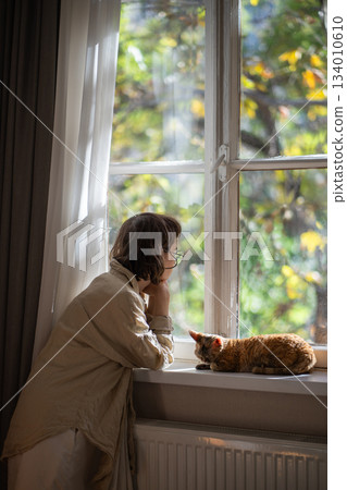 Melancholic pet owner looking at window in depression while staying at home in sunny summer day 134010610