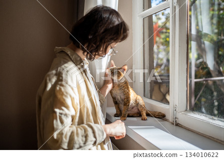 Pet lover caressing, stroking purring of pleasure beloved cat Devon Rex sitting on windowsill 134010622