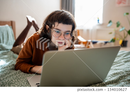 Focused teen girl studying, watching online lecture, webinar, seminar at laptop lying in bed at home 134010627