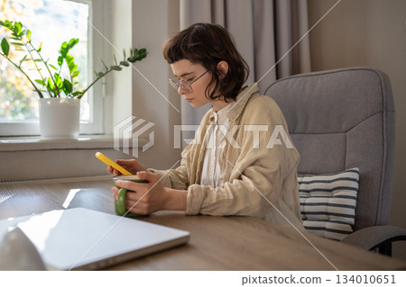 Focused girl freelancer browsing social networks looking in smartphone screen holding cup at home. Focused girl freelancer browsing social networks looking in smartphone screen holding cup at home. 134010651