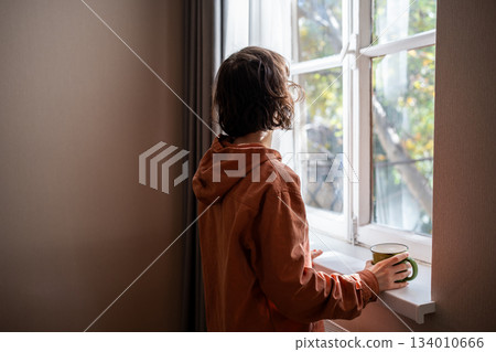 Woman looking at window drinking tea coffee on kitchen at home, rear view. Woman looking at window drinking tea coffee on kitchen at home, rear view. 134010666