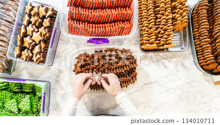 Hands carefully decorating a gingerbread man cookie amidst a festive spread of freshly baked holiday treats. Various cookie designs, including chocolate-dipped stars and red sugar-dusted cookies, are 134010711